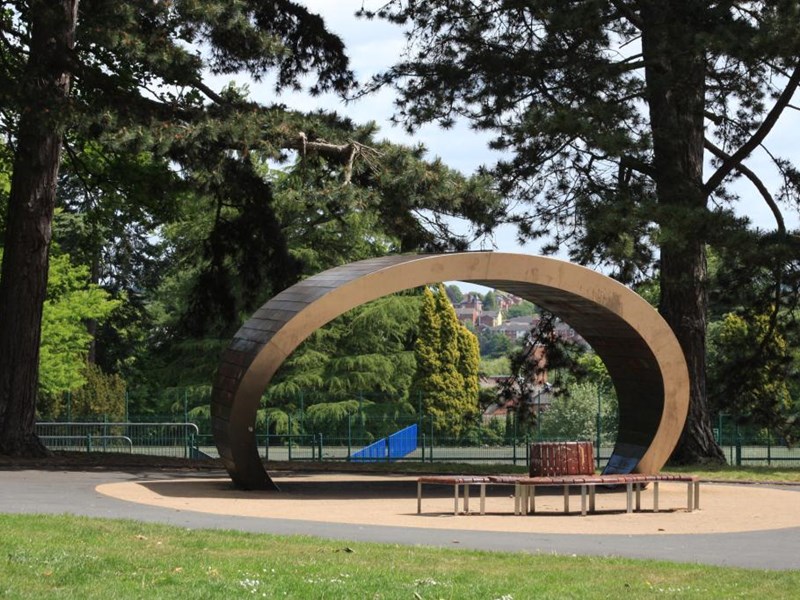 Curved structure with benches underneath forming shelter, skate park in distance.
